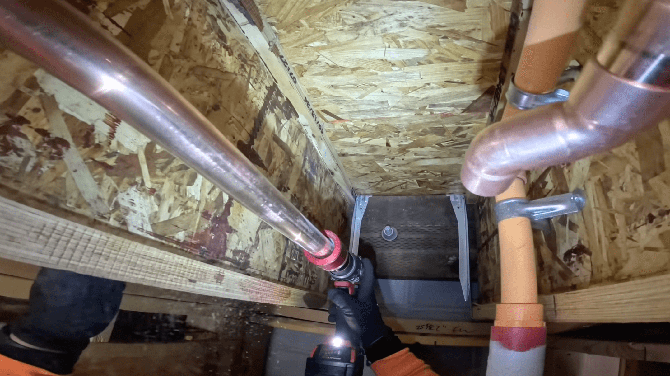 First-person GoPro view: a plumber's gloved hand pressing a copper pipe fitting up between floor joists, lit by a tool light.
